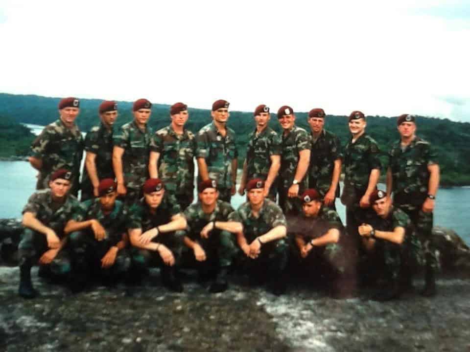 Group photo of military personnel in camouflage uniforms and berets, posing outdoors in front of a scenic landscape.